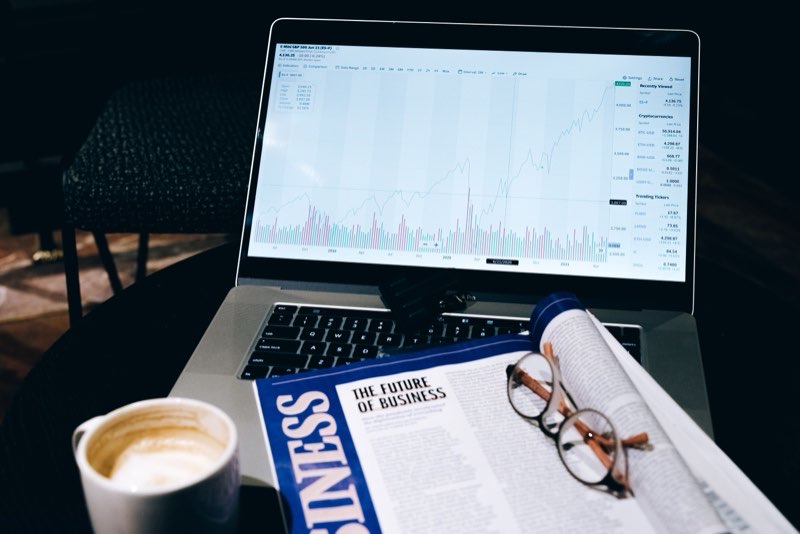 Laptop showing a market chart beside a business newspaper and reading glasses